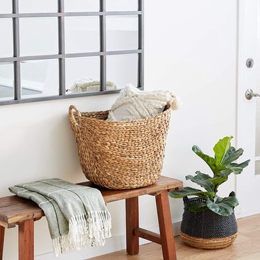 seagrass basket filled with a pillow on a bench with a throw and a potted plant on the floor