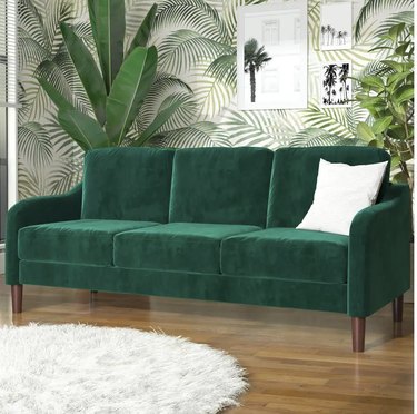 green velvet couch in a wallpapered room with palm trees sitting on a wood floor with a white area rug