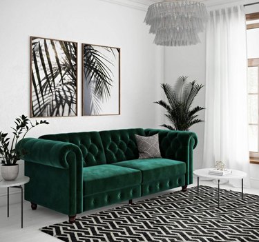 Green velevet chesterfield couch in white room with a black and white rug. Two plants sit on either side of the couch