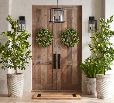Image of double wooden front doors with faux lemon and magnolia wreaths on either side. There are planters with lemon trees on either side of the doorway, and a entryway outdoor rug.
