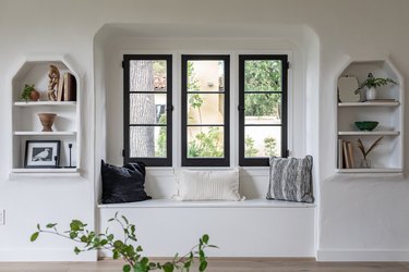 Three skinny windows are painted with black trim above a built-in bench and built-in bookshelves
