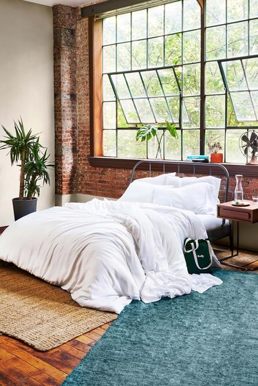 A fluffy white comforter in a bedroom with large windows and rugs.