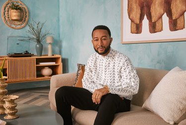 Musician John Legend sits on a couch wearing black pants and a white and black button-up shirt.
