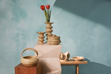 An assortment of decorative baskets on display in front of a blue wall, including woven flower vases, a small woven trinket dish and baskets with handles and lids.
