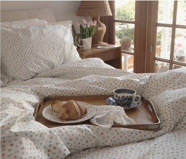 Breakfast tray with coffee and croissant on a bed with white cottagecore bedding with small flowers.