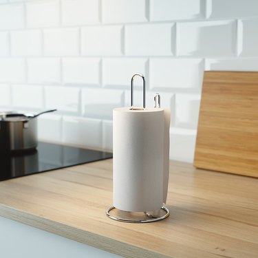 A silver paper towel holder on a light wood countertop in front of a white tile backsplash.