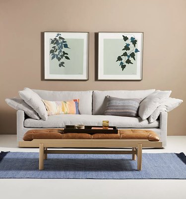 low coffee table with tufted leather top in front of light gray couch with pillows. The room is peach and there are two pieces of wall art with leaves.