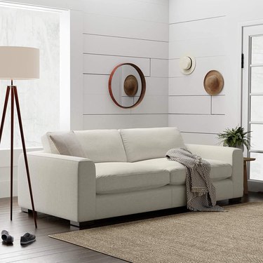 Beige couch in room with wooded floors, white walls, beige rug. There is a tripod lamp with a white shade in the corner