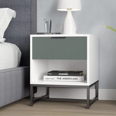 Image of white cuby style nighstand next to a gray bed with white linens. The nightstand is accessorized with books, a lamp and a glass of water.