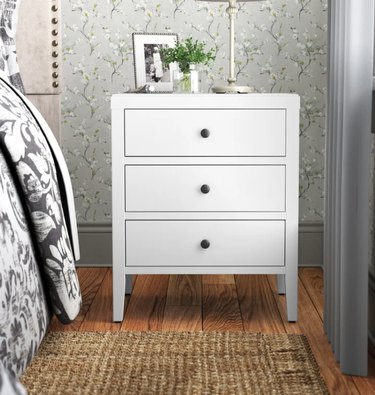Image of white nightstand in a room with wood floors and a jute rug. The walls are covered with floral wallpaper and there's a gray curtain in the room.
