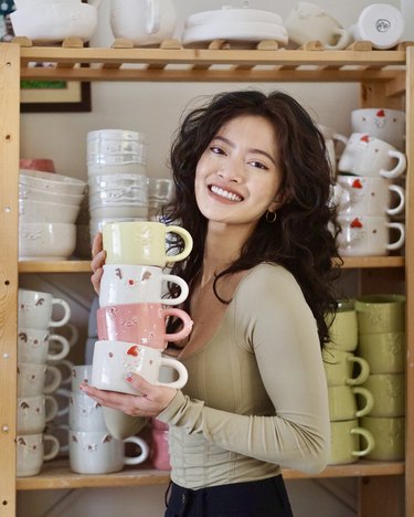 person with black hair and neutral long sleeve shirt holding a stack of colorful mugs, with shelves holding mugs in the background