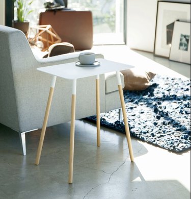 Image of white side table with wooden legs next to a beige couch. There's a blue rug in the room.
