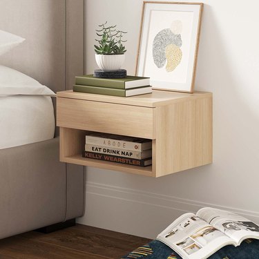 Image of a floating night stand in a light oak color. There are books, a plant, and a framed drawing on the nightstand.