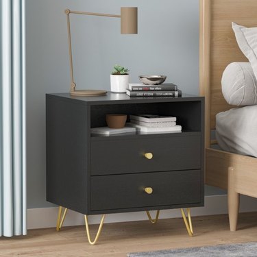 Image of 2-drawer black nightstand with gold accents. The nighstand is accessorized with a lamp, books and a plant.