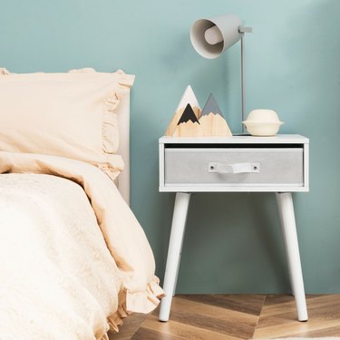 Image of white mid century nightstand  next to a bed with cream colored linens in front of a seagreen wall. The nightstand is accessorized with decorative items and a lamp.
