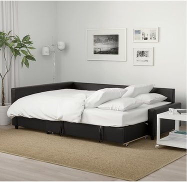 Image of Friheten pull-out sofa bed from Ikea, with white bedding, ready for guests. The sofa bed is in a white room, there's a jute rug underneath the couch and there are photos on the wall, a plant on the corner and a small white end table on the side.