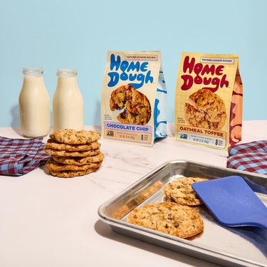 Bags of Home Dough's chocolate chip and oatmeal toffee cookie dough. There are surrounded by two glasses of milk, a tray of fresh-baked cookies, and blue and red gingham napkins.