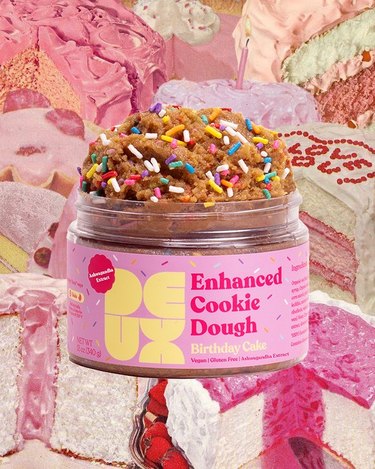 Deux's enhanced cookie dough in the birthday cake flavor. The jar is a Barbie shade of pink and there are rainbow sprinkles on top of the dough. In the background, there is a collage of pink cakes.
