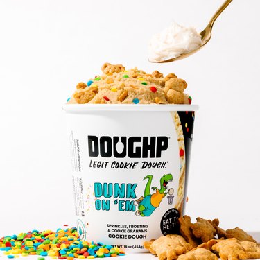 Doughp's "dunk on em" cookie dough pint surrounded by teddy grahams and rainbow sprinkles. A spoon with frosting on it is about to dip into the dough.