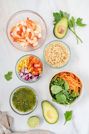 Bowls filled with the chimichurri sauce, pink shrimp, rice, carrots, spinach, cherry tomatoes, corn, and red onion. The bowls are surrounded by avocado halves, a lime half, and cilantro stems.