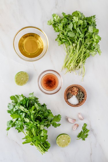 A bundle of cilantro, parsley, garlic cloves, half a lime, and small bowls holding red winder vinegar, olive oil, and salt, black pepper, dried oregano, and red pepper flakes. All have been placed on a marble countertop.