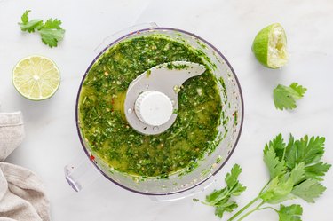 A food processor bowl filled with the chimichurri sauce. It is surrounded by limes and cilantro.