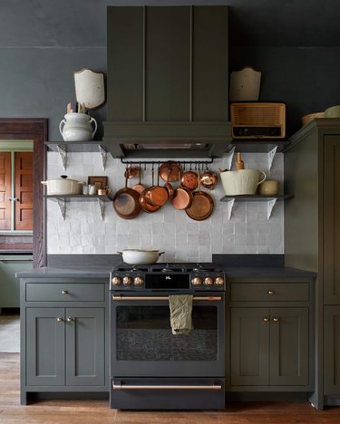 Kitchen with dark green cabinets, white zelig backsplash, pot rack, copper pots, wood floor, dark gray counters.