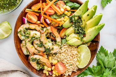 A wood bowl filled with chimichurri shrimp. rice, avocado slices, lime, and a mix of cherry tomatoes, corn, and carrots. On the surrounding marble countertop, there is a lime wedge, bundle of cilantro, and bowl of chimichurri sauce.
