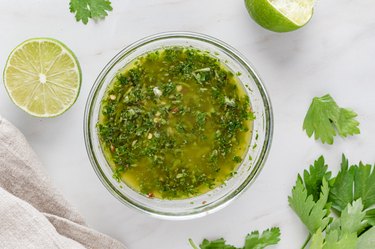 The chimichurri sauce in a clear glass bowl, still surrounded by two lime halves and cilantro.