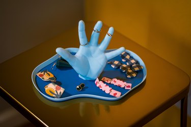 A close up of a decorative tray on a beige table. The tray is bright blue  with a squiggly base that's shaped like a body of water; in the middle is a sculpture of a life-sized hand that is reaching out. Gold rings are placed on three fingers of the sculpture, and an assortment of color earrings are placed on the flat surface of the tray.