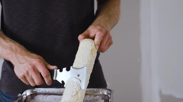 A person using a multipurpose painting tool to scrape excess paint off of a paint roller.