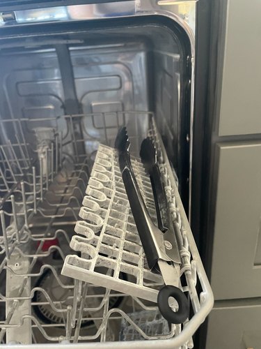 Inside of a dishwasher, top bracket raised, with a pair of tongs resting on the flap