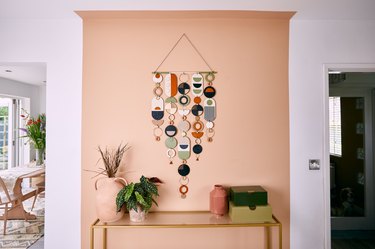 Hanging wall piece on a peach colored wall with a table topped with plants and boxes