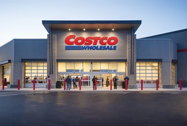 A Costco warehouse at night. The red and blue logo is on the front of the building, with four lights above it. There is a line of people outside the doors, spilling onto the sidewalk.