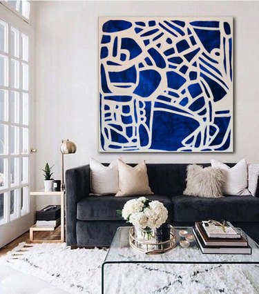 Blue and white painting on a white wall in a room with a navy blue couch, clear coffee table, and white rug