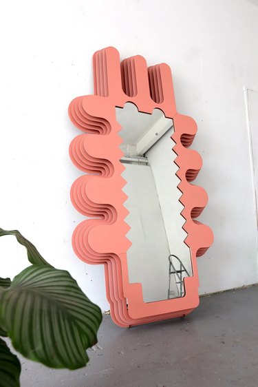 Mirror with a salmon-colored border leaning against a white wall. There is a plant in the right corner.
