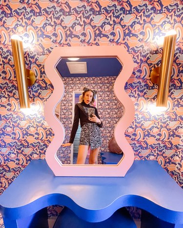 A vanity in the Trixie Motel featuring a wavy mirror lined in pink on a rich blue wavy table. In the background, the wallpaper shows wavy Saturn-like planets in pink and blue.