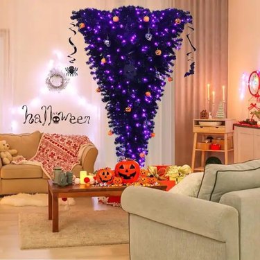 An upside-down Halloween tree in a living room. The tree features purple lights along with both pumpkin and skull ornaments. There are two cream-colored sofas in the living room and a coffee table topped with pumpkin bowls.
