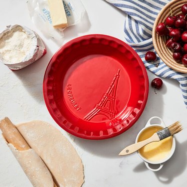 Red pie dish from above next to a bowl of cherries, stick of butter, flour, and rolling pin