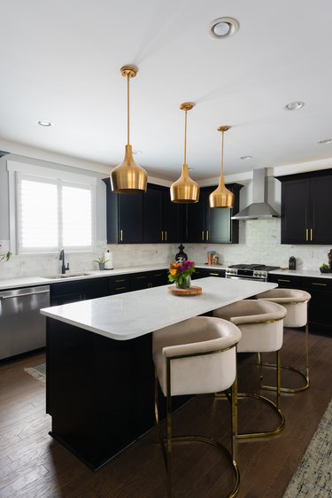 Kitchen with a white and wooden island, cream colored bar chairs, brass hanging lamps. white countertops, and white walls