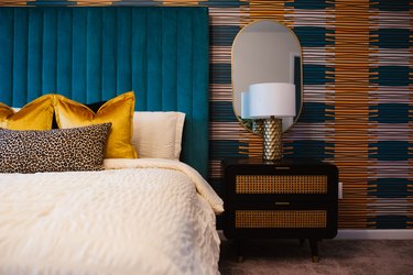 Bedroom with cream colored bedding, mustard yellow pillows, a blue/green headboard, side table with a lamp and mirror