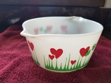 The Lucky in Love Pyrex dish on a red blanket. The dish is a translucent white and features red hearts, green clovers, and green grass spikes at the bottom.
