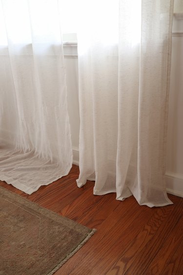 One white curtain panel that is hemmed hanging next to another white curtain panel that is too long
