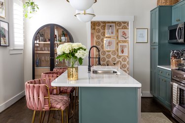 Kitchen with white and green island, pink bar chairs, a brass colored vase with white flowers, white walls