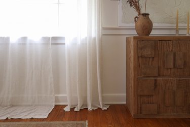 One white curtain panel that is hemmed hanging next to another white curtain panel that is too long