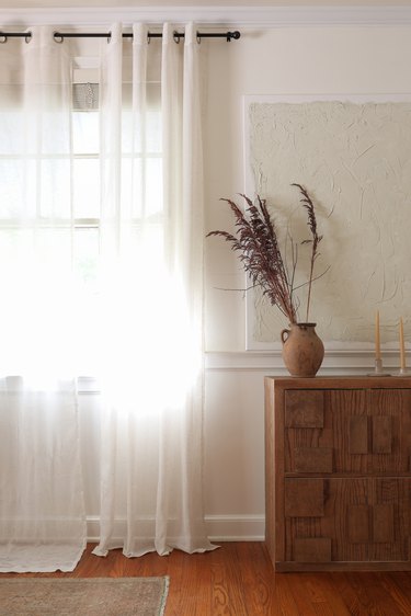 One white curtain panel that is hemmed hanging next to another white curtain panel that is too long