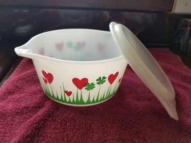 The Lucky in Love Pyrex dish on a red blanket. The dish is a translucent white and features red hearts, green clovers, and green grass spikes at the bottom. There is also a white lid leaning on the right side of the dish.