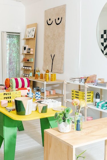 Photo of  a corner of the home decor shop YOWIE. There is a long window in the background, next to it is a wooden shelf with various items. A beige shag rug showing a minimal line drawing of a nude person hangs on the wall next to the bookshelf. Below the rug are 3-tiered tables filled with books, plates, and other decor items. In the middle of the space is a yellow table with green legs filled with items like green and beige planters, a white and red towel, and more books.