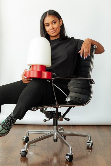 Photo of Shannon Maldonado, a black woman with straight brown hair that falls to her shoulders. She is wearing a black shirt tucked into black pants, and dark green and black sneakers with grey socks. She sits playfully on a swivel chair, one hand rest on the top of the chair, while her right arm holds a bulbous white and red lamp.