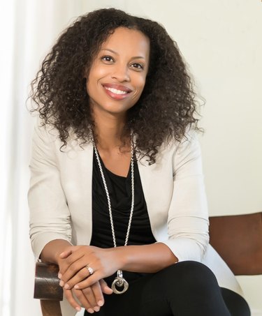 A photo of Laura Hodges, a black woman with shoulder-length curly hair. She is wearing a white blazer, black T-shirt, and black pants, along with a long silver necklace with a thick silver circle at the end.